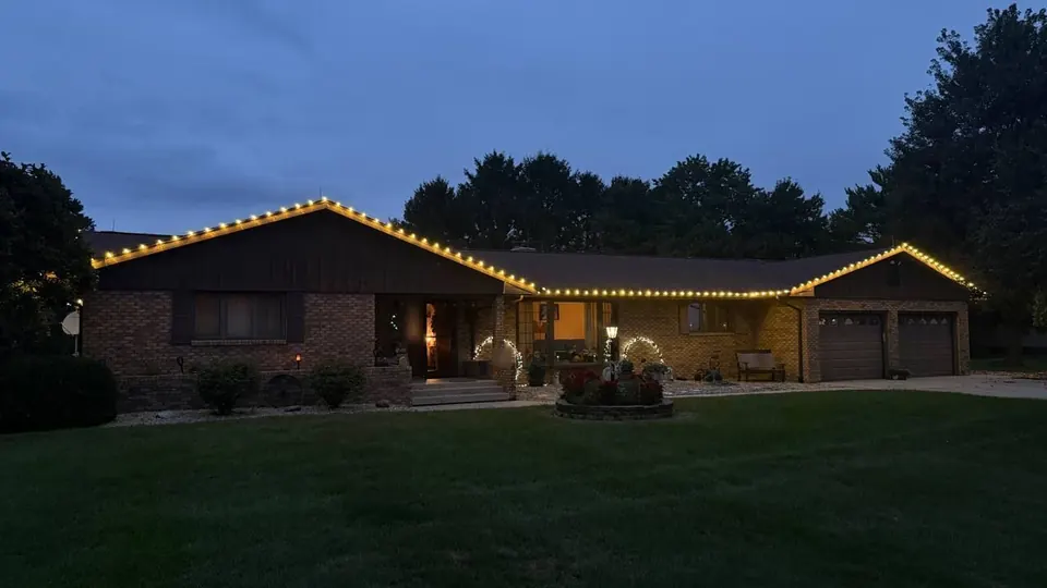 Brick ranch home at dusk with warm white Christmas lights outlining the roofline and gables
